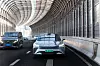 Modernes Elektrofahrzeug eines chinesischen Herstellers fährt auf einer mehrspurigen Schnellstraße durch eine futuristisch überdachte Tunnelkonstruktion mit geschwungener Stahl-Glas-Architektur; das Fahrzeug ist mit Sensorik für automatisiertes Fahren ausgestattet und bewegt sich im fließenden Verkehr neben weiteren Pkw, was die zunehmende Verbreitung von Fahrerassistenz- und autonomen Fahrfunktionen im urbanen Umfeld Chinas symbolisiert.