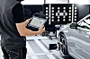 Eine Person in Werkstattkleidung hält ein robustes Tablet vor einem grauen Fahrzeug. Im Hintergrund steht ein Kalibrieraufbau mit schwarz-weißen Markierungstafeln. Am Vorderrad des Autos ist Messzubehör angebracht. Die Szene zeigt eine technische Prüfung oder Kalibrierung von Fahrzeugsensoren in einer hellen Werkstattumgebung.