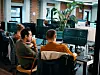Mehrere Personen arbeiten in einem offenen Büro an Computern. Auf mehreren Bildschirmen sind Codezeilen und Entwicklungsumgebungen zu sehen. Im Vordergrund sitzen zwei Mitarbeitende an einem Schreibtisch, im Hintergrund weitere Arbeitsplätze. Das Büro wirkt modern und hell, mit großen Pflanzen und Backsteinwänden.