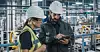 CSP: Zwei Beschäftigte prüfen Produktionsdaten an einem Tablet in einer modernen Fertigungsanlage mit Maschinen und Fördertechnik im Hintergrund.