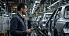Mitarbeiter mit Tablet in einer Autofabrik neben einer Reihe montierter Fahrzeuge.