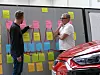 Zwei Männer besprechen Haftnotizen an einem Board, vorne ist ein rotes Auto zu sehen.