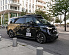 Schwarzer, autonom fahrender Volkswagen ID. Buzz von Moia im Hamburger Stadtverkehr, aufgenommen am Straßenrand vor einem modernen weißen Wohn- und Bürogebäude mit Balkonen, großen Fensterflächen und jungen Straßenbäumen. Auf dem Fahrzeug sind deutlich die Volkswagen-Logos, die Aufschrift „Self-Driving“, der Moia-Schriftzug sowie Hinweise auf den autonomen Testbetrieb zu erkennen. Auf dem Dach befindet sich ein großer Sensoraufbau für das selbstfahrende System, ergänzt durch weitere Kameras und Sensoren rund um das Fahrzeug. Der vollelektrische Shuttle steht exemplarisch für den im Juli 2026 gestarteten Fahrgast-Testbetrieb von Moia in Hamburg: Vorab registrierte Nutzerinnen und Nutzer können autonome Fahrten über die Moia-App buchen, später soll dies auch über hvv switch möglich sein. Im Rahmen des Projekts ALIKE sollen bis zu zehn autonome ID. Buzz in Teilen von Winterhude, Barmbek und Wandsbek eingesetzt werden; während der Testphase bleiben Sicherheitsfahrerinnen oder Sicherheitsfahrer an Bord.