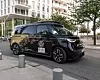 Schwarzer, autonom fahrender Volkswagen ID. Buzz von Moia im Hamburger Stadtverkehr, aufgenommen am Straßenrand vor einem modernen weißen Wohn- und Bürogebäude mit Balkonen, großen Fensterflächen und jungen Straßenbäumen. Auf dem Fahrzeug sind deutlich die Volkswagen-Logos, die Aufschrift „Self-Driving“, der Moia-Schriftzug sowie Hinweise auf den autonomen Testbetrieb zu erkennen. Auf dem Dach befindet sich ein großer Sensoraufbau für das selbstfahrende System, ergänzt durch weitere Kameras und Sensoren rund um das Fahrzeug. Der vollelektrische Shuttle steht exemplarisch für den im Juli 2026 gestarteten Fahrgast-Testbetrieb von Moia in Hamburg: Vorab registrierte Nutzerinnen und Nutzer können autonome Fahrten über die Moia-App buchen, später soll dies auch über hvv switch möglich sein. Im Rahmen des Projekts ALIKE sollen bis zu zehn autonome ID. Buzz in Teilen von Winterhude, Barmbek und Wandsbek eingesetzt werden; während der Testphase bleiben Sicherheitsfahrerinnen oder Sicherheitsfahrer an Bord.