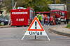 Ein Warndreieck mit der Aufschrift „Unfall“ steht auf einer Straße. Im unscharfen Hintergrund befinden sich mehrere Feuerwehrfahrzeuge und Einsatzkräfte.
