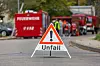 Ein Warndreieck mit der Aufschrift „Unfall“ steht auf einer Straße. Im unscharfen Hintergrund befinden sich mehrere Feuerwehrfahrzeuge und Einsatzkräfte.