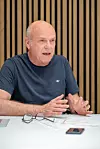 CIO Loydl sitzt an einem Tisch mit Unterlagen und spricht gestikulierend vor Holzpaneel-Hintergrund.