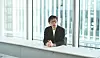Shizuka Hirakawa sitzt in einem hellen, modern verglasten Büro an einem langen Tisch. Er trägt ein dunkles Sakko und eine Brille und spricht mit gefalteten Händen in Richtung Kamera.