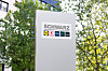 Weißes Schild mit Schwarz-Logo vor Bäumen im Freien.