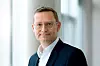 Porträt eines Managers mit Brille im dunklen Anzug und weißen Hemd vor einem hellen, unscharfen Bürohintergrund.