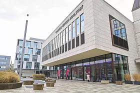 Modernes Gebäude mit Glasfront, gepflastertem Vorplatz und runden Sitzbänken im Außenbereich.