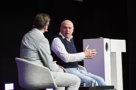 Zwei Männer auf einer Bühne, einer spricht und gestikuliert neben einem weißen Podium.