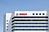 Außenansicht eines modernen Bosch-Gebäudes mit weißer Fassade, dunklen Fensterbändern und blauem Himmel im Hintergrund. Im oberen Bereich der Front ist das Bosch-Logo in Rot zusammen mit dem runden Markensymbol angebracht. Das Bild steht für die angekündigte Neuordnung der Bosch-Konzernstruktur in Deutschland, bei der der Bereich Mobility sowie Bosch Digital, Global Business Services und Bosch Real Estate bis 2027 als eigenständige Tochtergesellschaften innerhalb der Bosch-Gruppe aufgestellt werden sollen.
