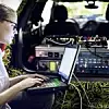Eine Frau, Mitarbeiterin von BMW, sitzt mit ihrem Laptop vor dem Kofferraum eines Fahrzeugs, in dem elektronische Gerätschaften verkabelt sind.