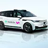 Weißes, autonom fahrendes Shuttle-Fahrzeug mit schwarzem Dach und Sensoren auf dem Dach. An den Fahrzeugseiten sind die Logos von „Apollo Go“ und „Lyft“ zu sehen. Das Auto steht auf glänzendem Boden vor hellem Hintergrund.
