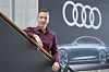 Ein Mann mit kurz geschnittenen Haaren und weinrotem Hemd steht an einem Treppengeländer. Im Hintergrund sind das Audi-Logo und die Abbildung eines futuristischen Autos auf einer Wand zu sehen.
