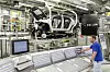 Ein Volkswagen-Mitarbeiter bedient eine Steuerkonsole in einer hochautomatisierten Fertigungslinie. Im Hintergrund wird eine Fahrzeugkarosserie von Robotern und Maschinen bewegt.