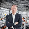 Joachim Franz steht mit verschränkten Armen. Im Hintergund ist eine hochmoderne Smart Factory mit automatisierten Fertigungsrobotern.