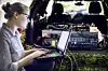 Eine Frau, Mitarbeiterin von BMW, sitzt mit ihrem Laptop vor dem Kofferraum eines Fahrzeugs, in dem elektronische Gerätschaften verkabelt sind.